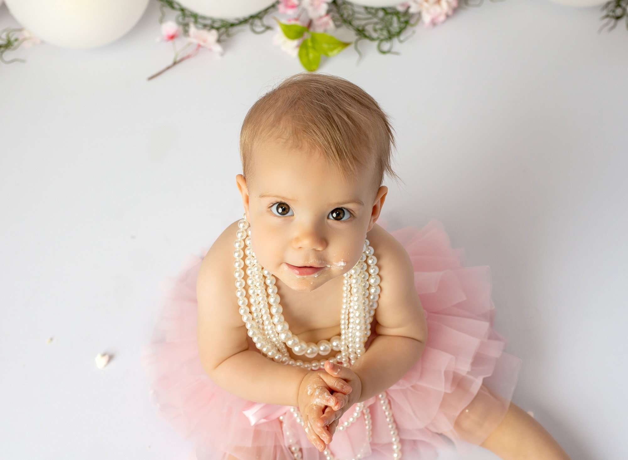 brown eyed baby girl wearing layered pearl necklace and pink tutu with cake frosting smeared on cheek and hands