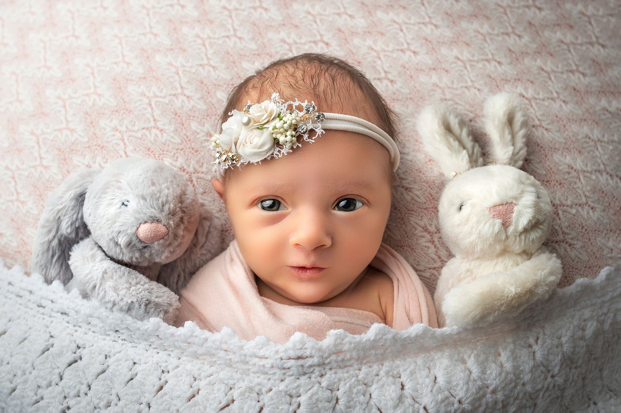 Awake newborn Baby cozily tucked in with two cuddly bunnies under a soft blanket, capturing a moment of pure innocence.