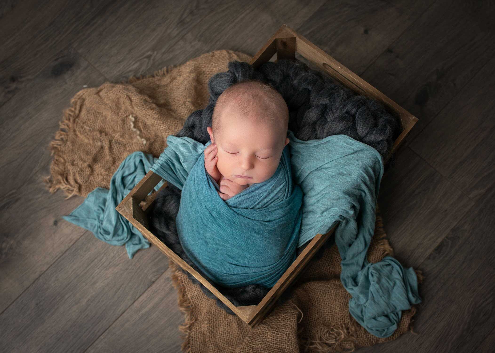 newborn boy swaddled in teal wrap sleeping in crate on grey blanket
