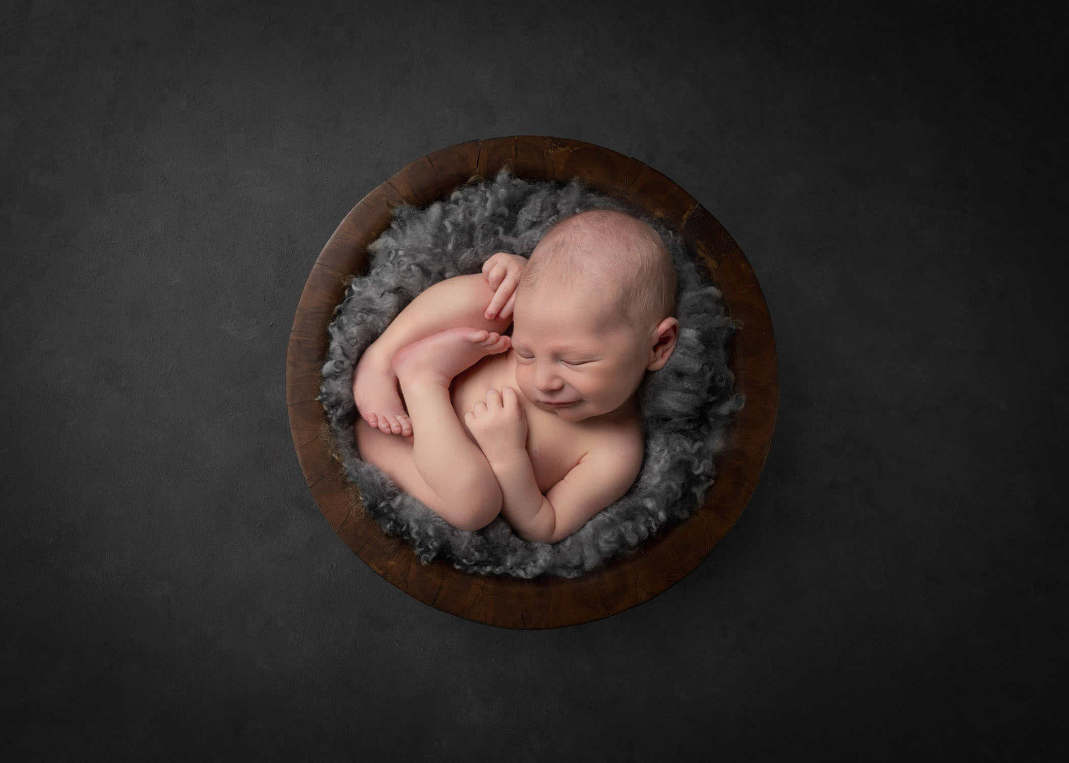 Newborn boy sleeping curled in bowl on grey fluff