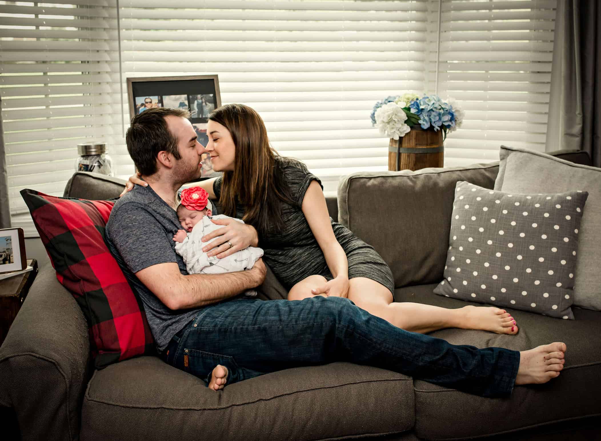 Mom and Dad about to kiss while holding sleeping newborn on couch