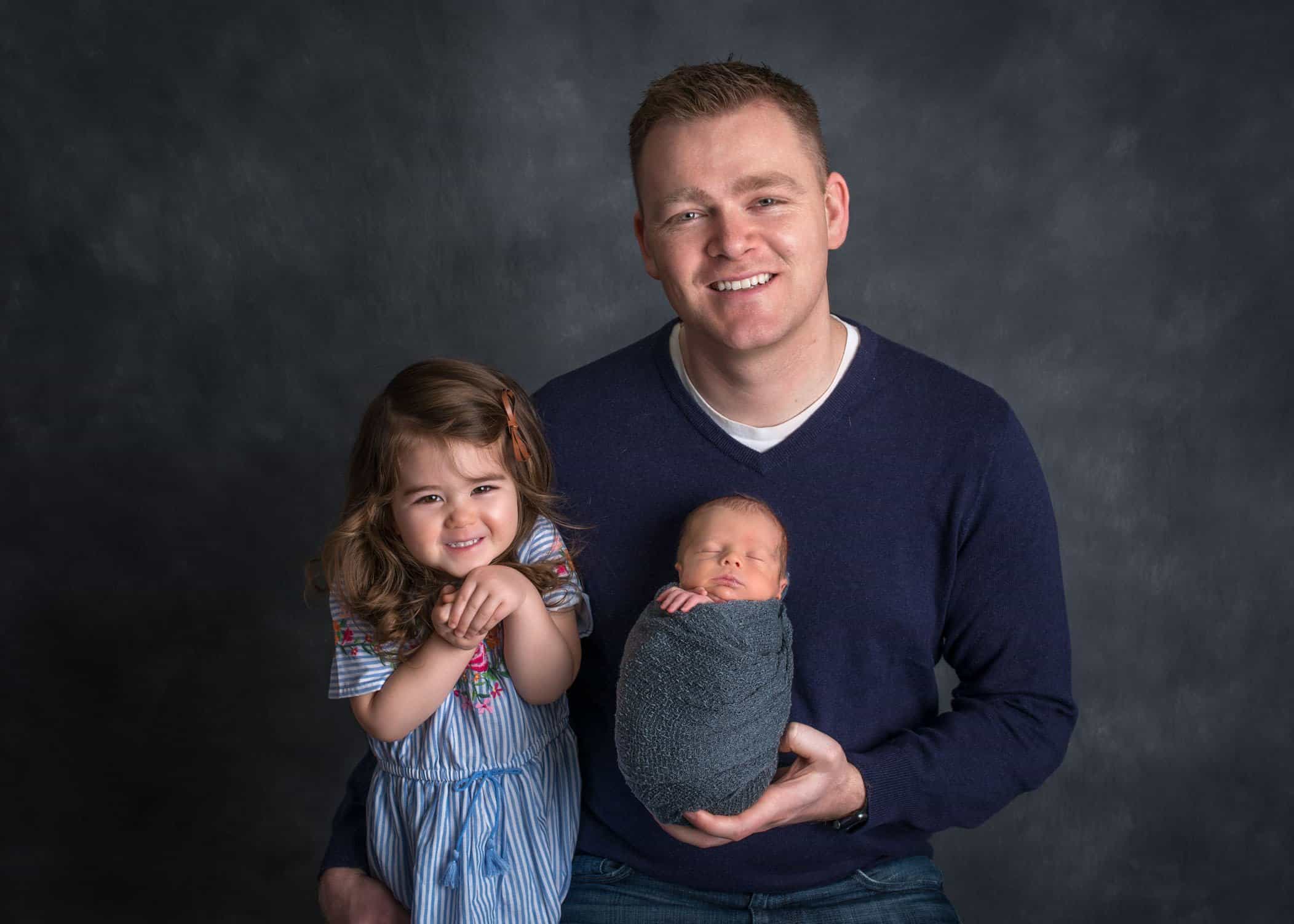 Dad holding newborn son in one hand and hugging toddler sister with other arm