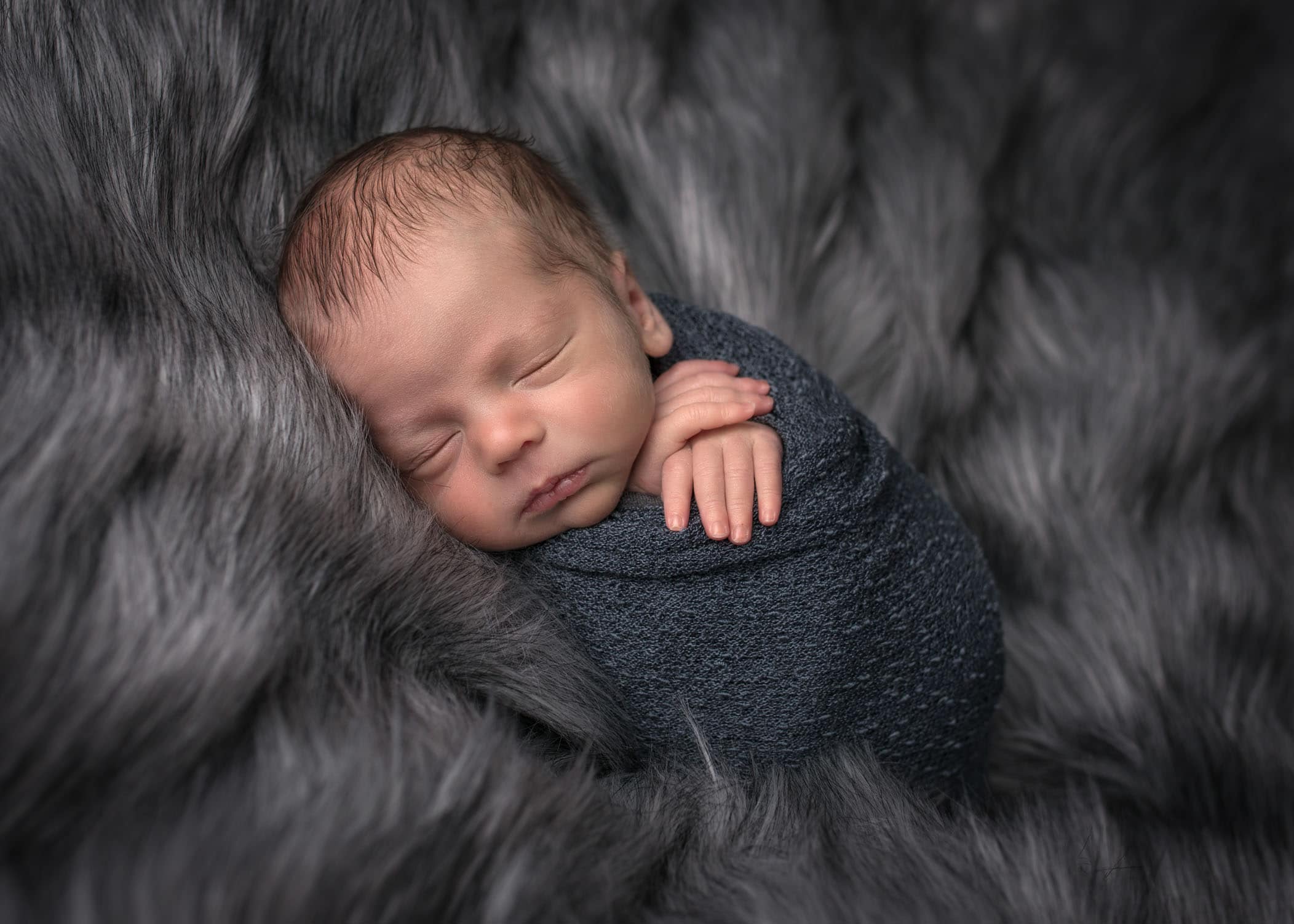 newborn boy sleeping with hands folded under his chin while swaddled and on grey fur