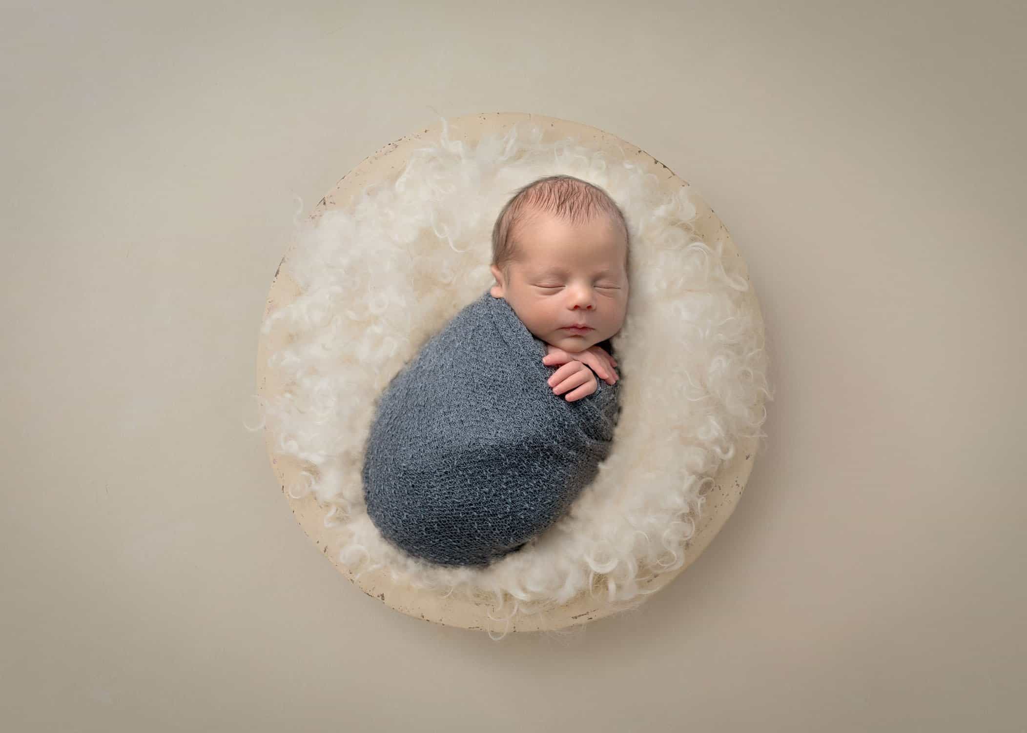 newborn boy swaddled and sleeping on cream fluff in cream wooden bowl