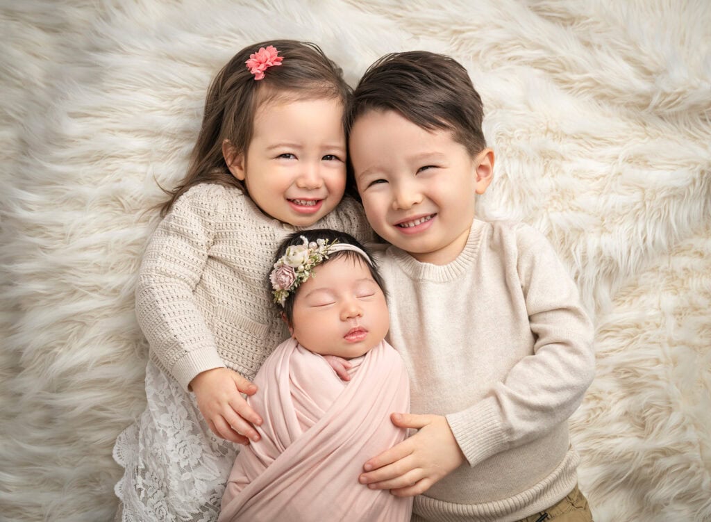 Siblings on ivory throw with swaddled newborn in blush wrap, soft natural smiles.