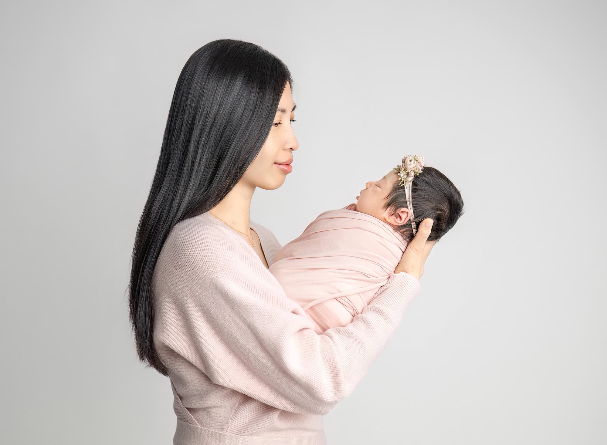Mother in pale pink holding wrapped newborn against clean light grey backdrop.