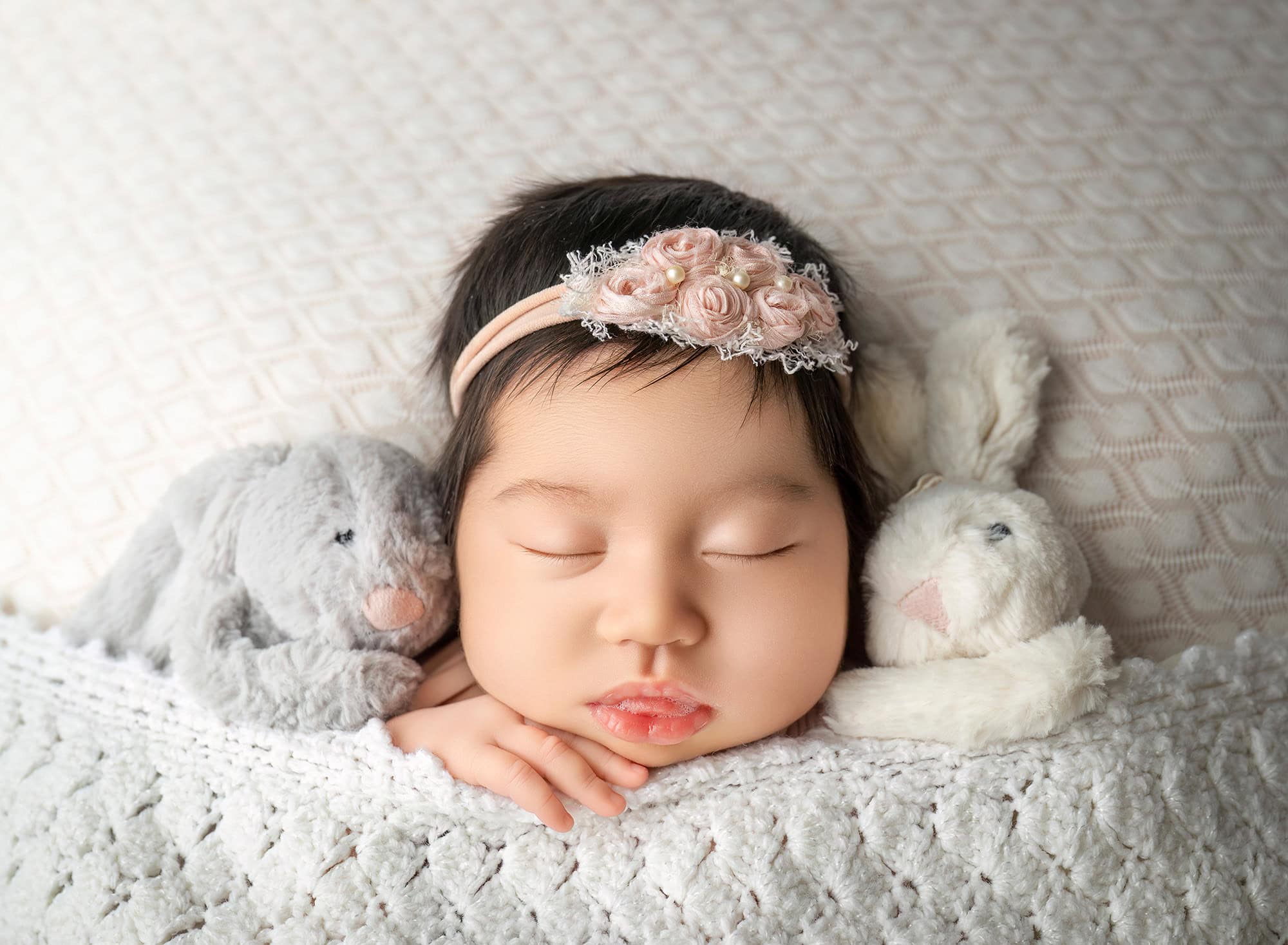 Close-up of sleeping newborn between two plush bunnies, textured white blanket.