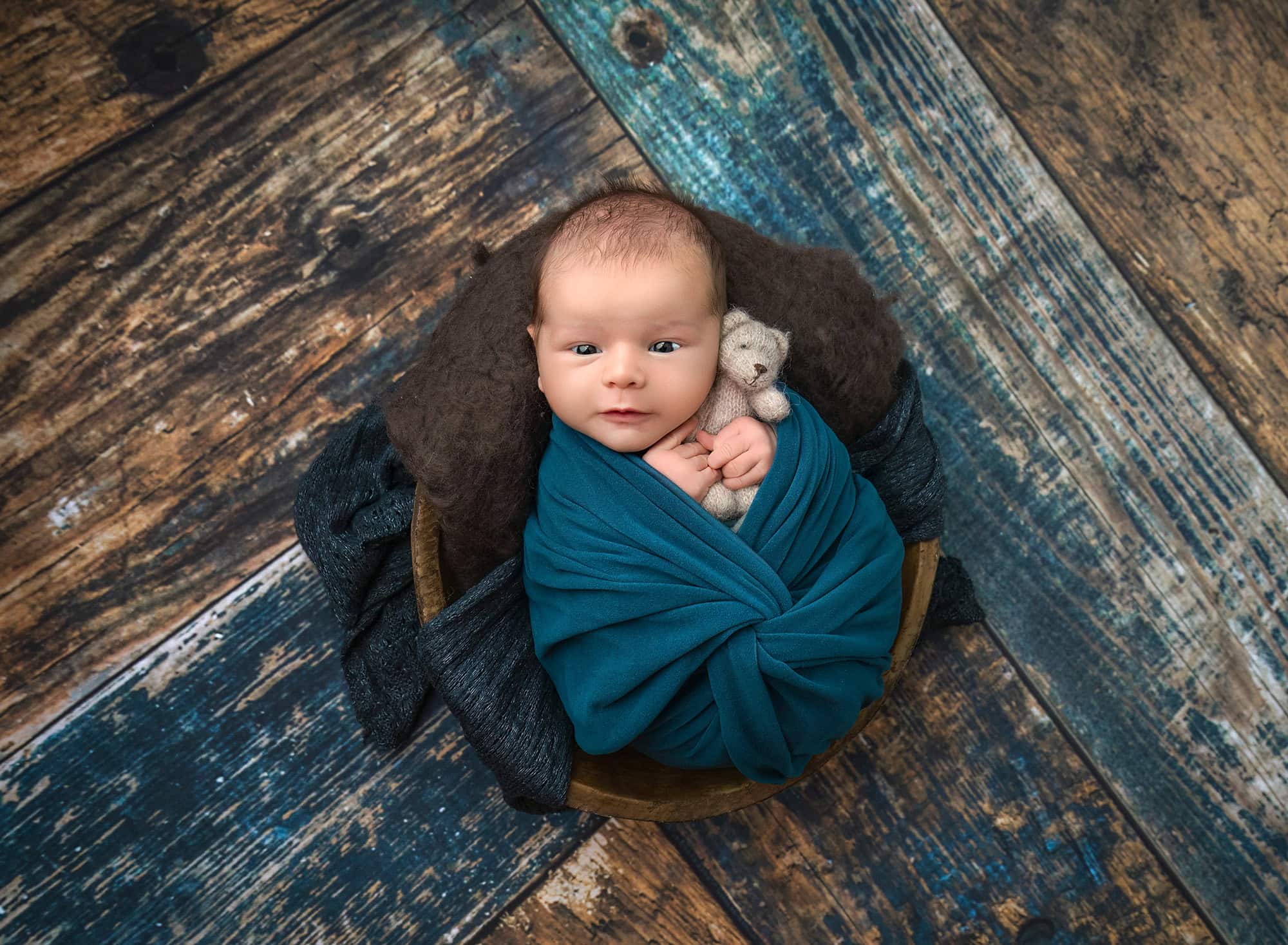 awake baby Leif swaddled in teal holding a tiny bear on rustic wood