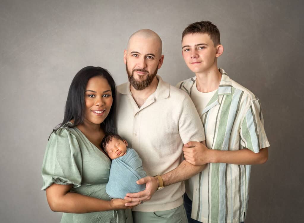 Studio family portrait with mom, dad, teen brother and swaddled newborn in sage against a soft gray backdrop.
