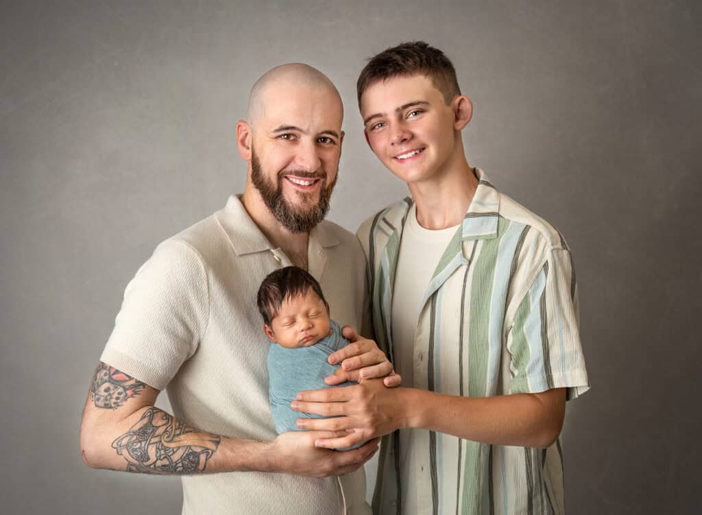 Dad and big brother smiling while safely holding a swaddled newborn in sage during a studio portrait.