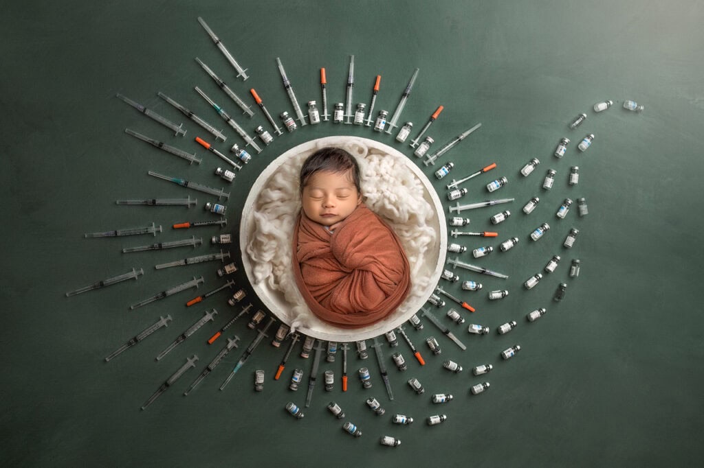 Sleeping newborn in rust wrap in a bowl, surrounded by IVF syringes and medication vials—fertility tribute portrait.