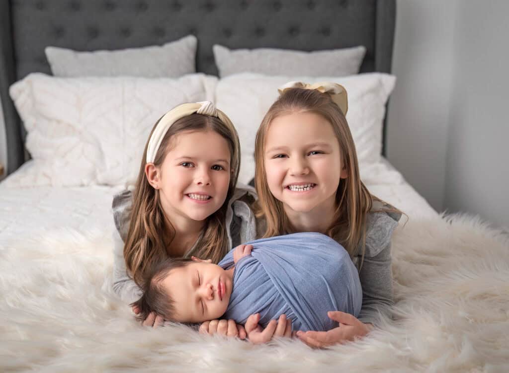 Siblings Vivian and Isabelle smiling while holding baby Max on a bed.