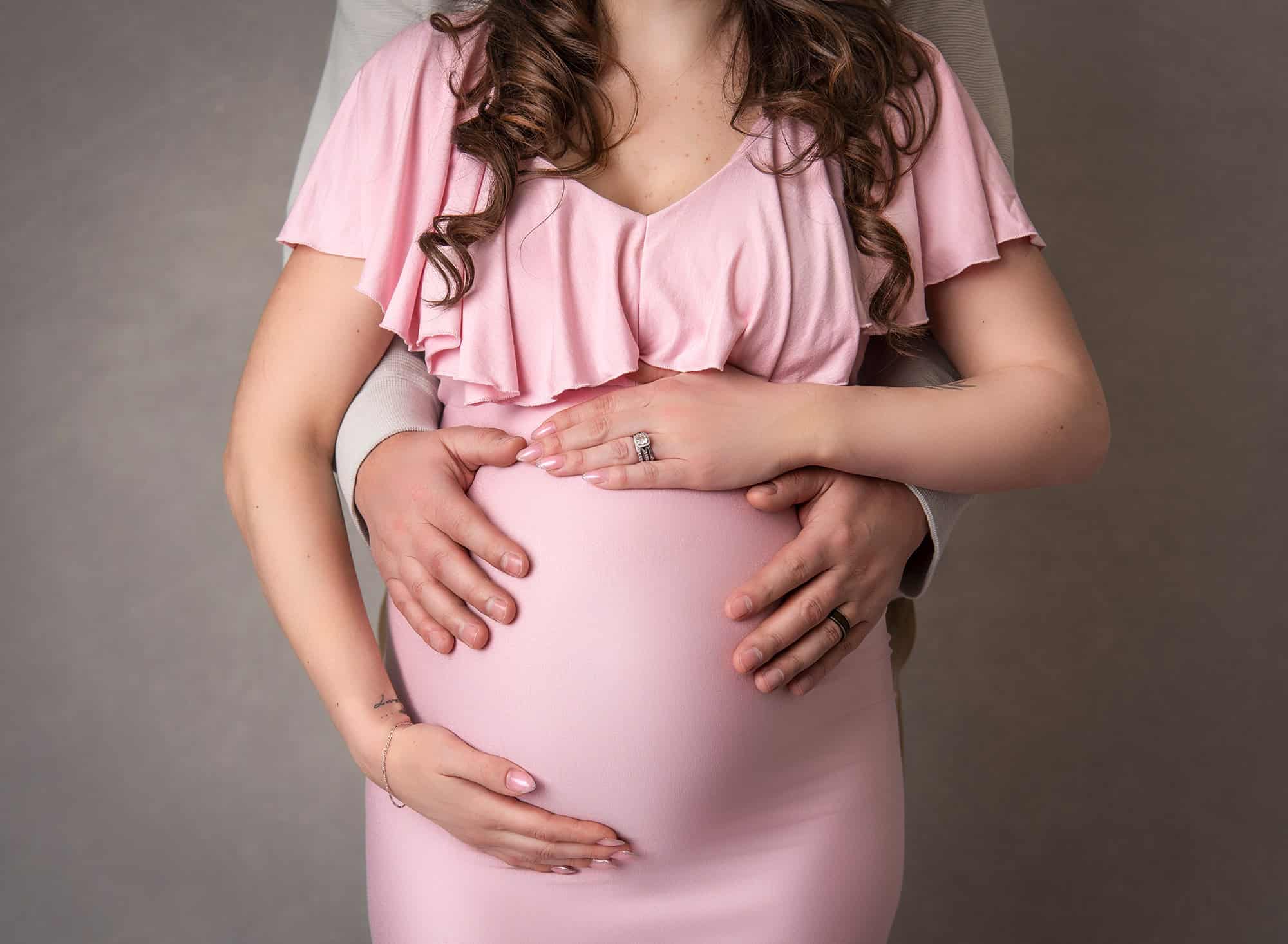 woman wearing baby pink maternity dress cradling growing belly with her and dads hands