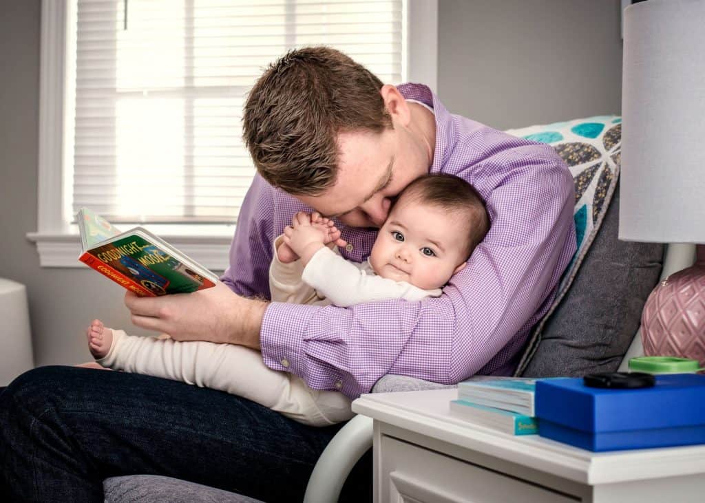 6 month old baby sitting with Daddy in the rocking chair sharing a book, some kisses and playing with her feet