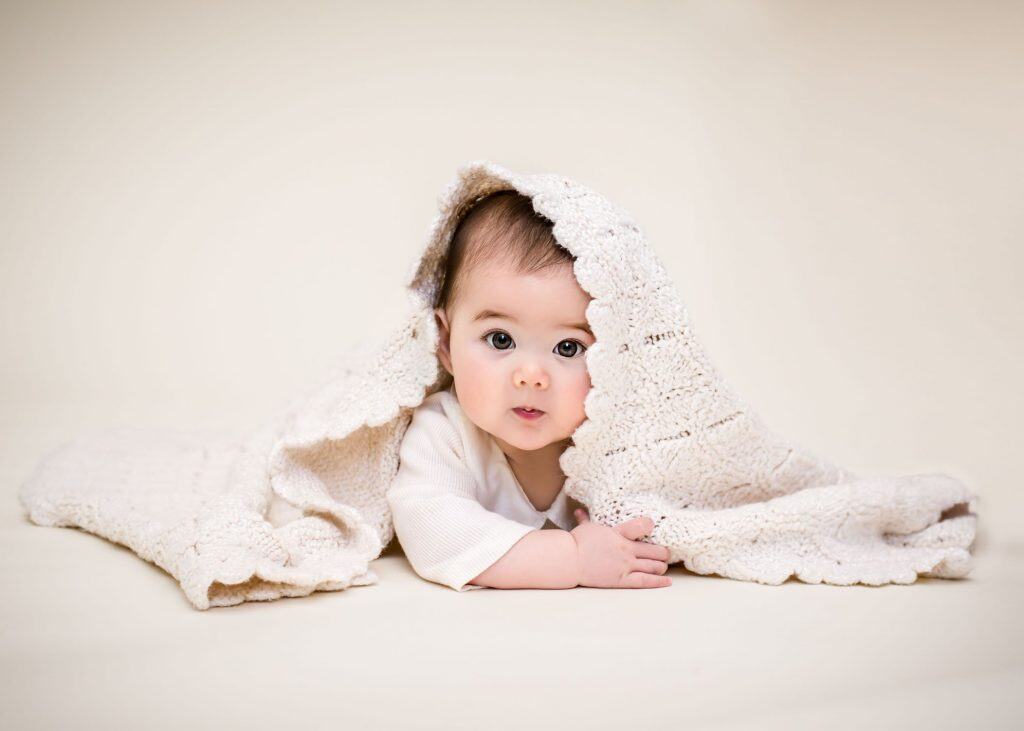 6 month old baby lying on her tummy playing peek a boo on a cream background