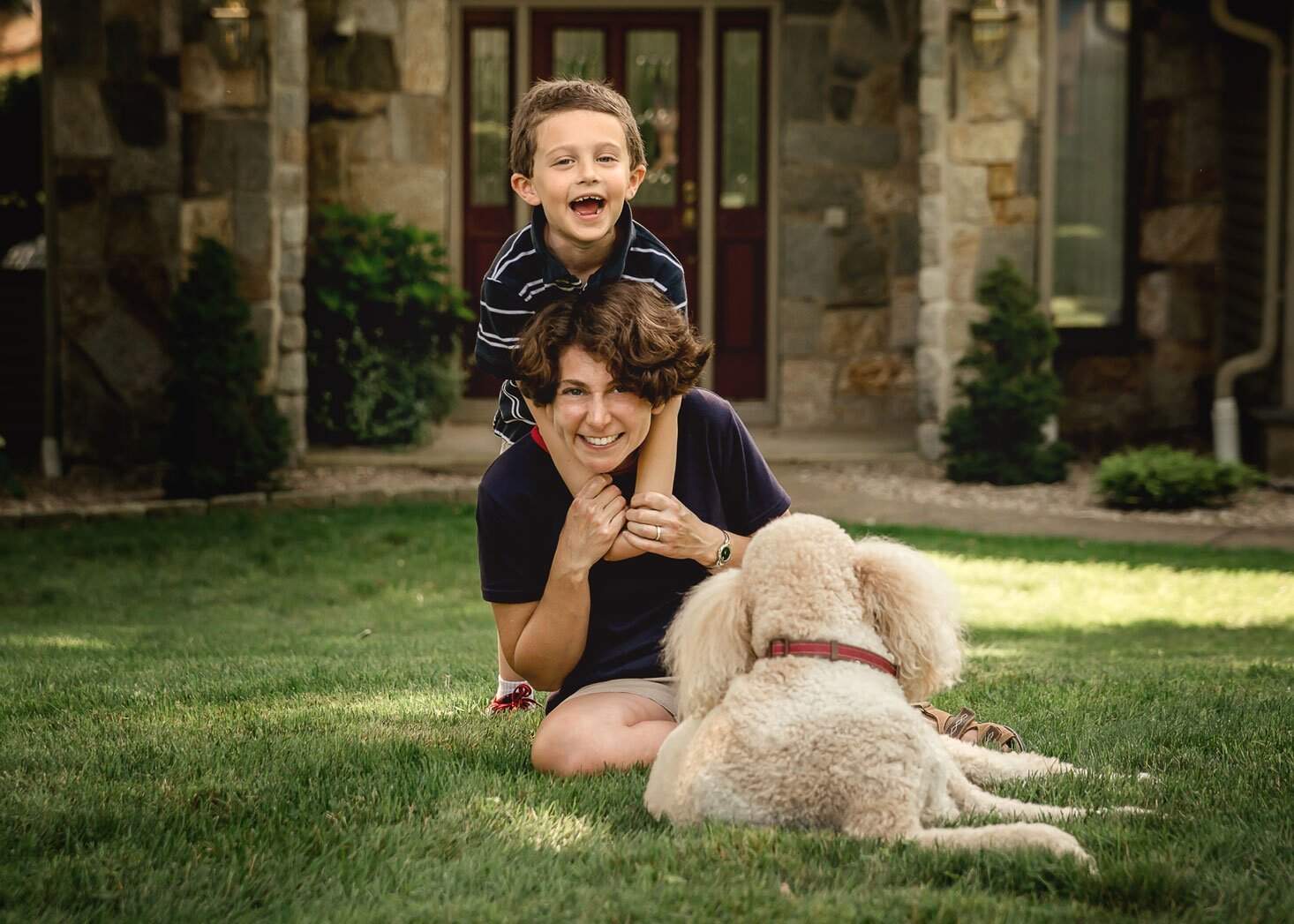 young son hugs mom from behind around her neck with dog outside in summer