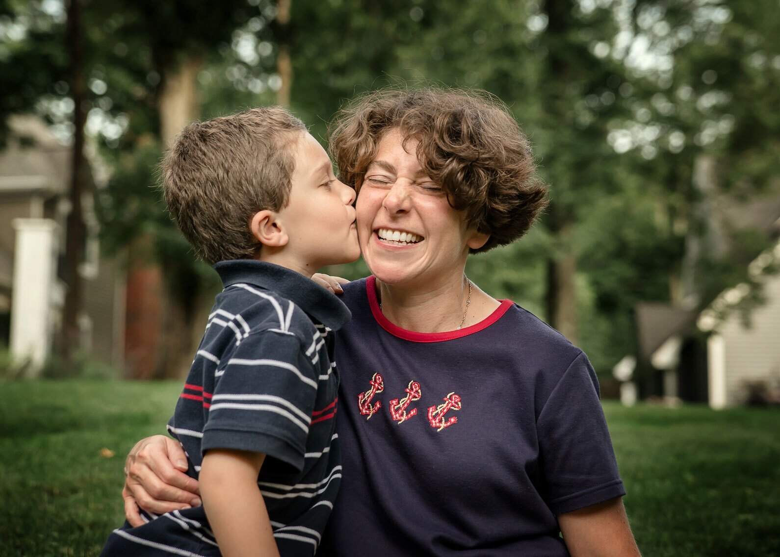 young son kisses laughing Mom on the cheek outside in summer