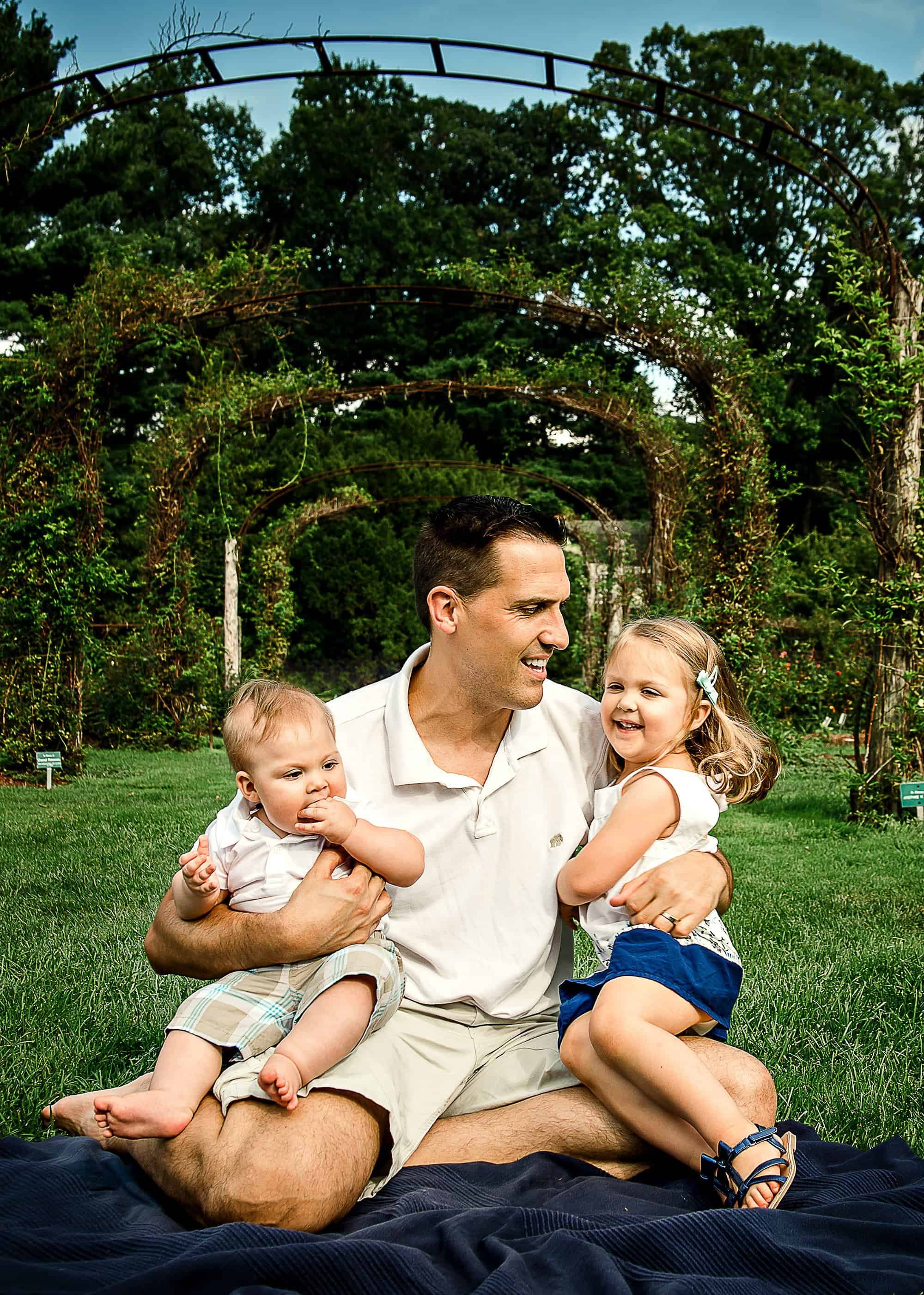 Daddy holding his 2 small children on his lap laughing in the garden