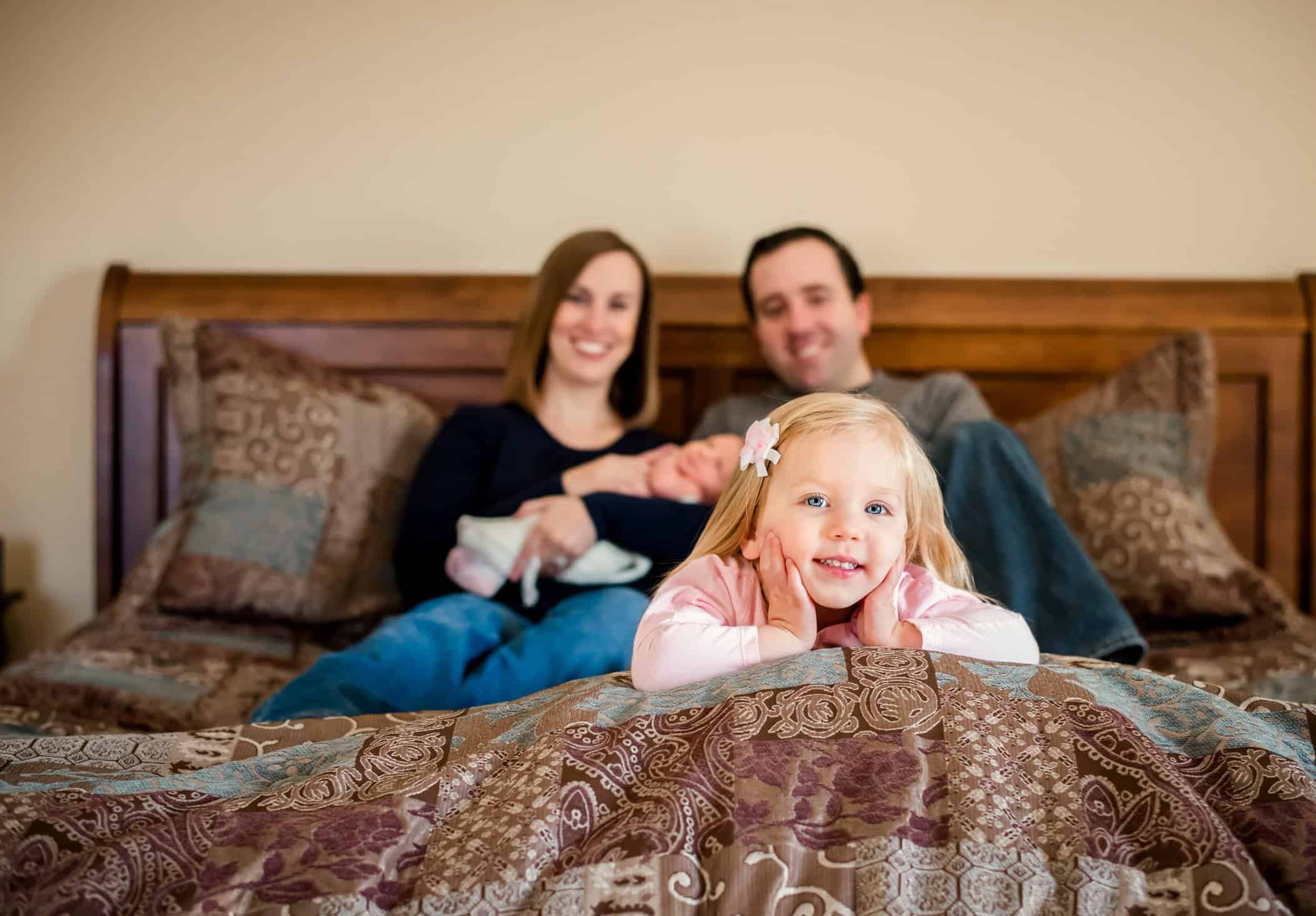 older sister lying on bed in front of Mom and Dad holding newborn brother