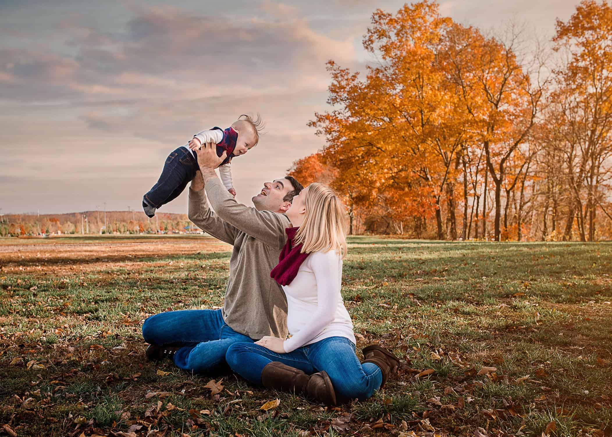 Daddy flying baby boy outside with fall foliage behind them