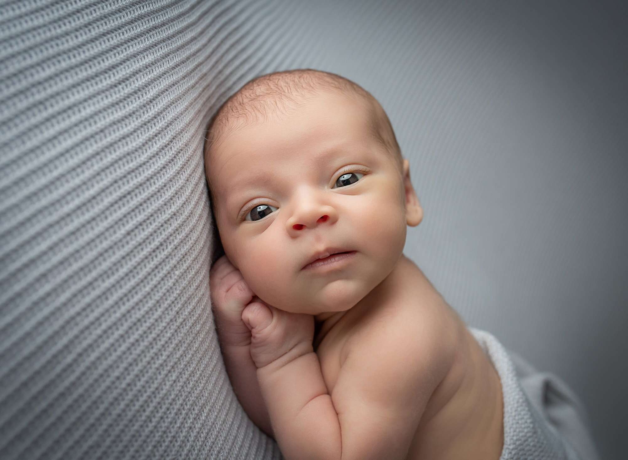 awake newborn baby boy looking at the camera as he lays on his side on a grey background best time to take newborn photos