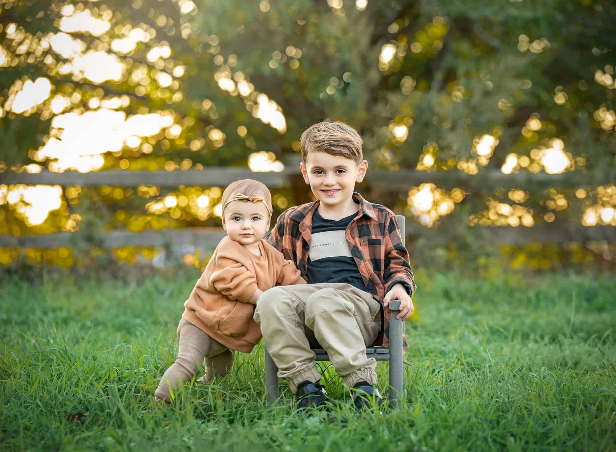 young boy sitting in nature with toddler sister standing next to him