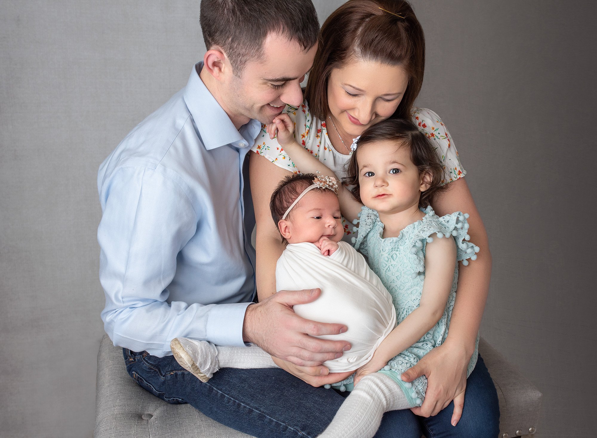 couple holding newborn baby girl swaddled in white with big sister in lace blue dress on dark gray backdrop