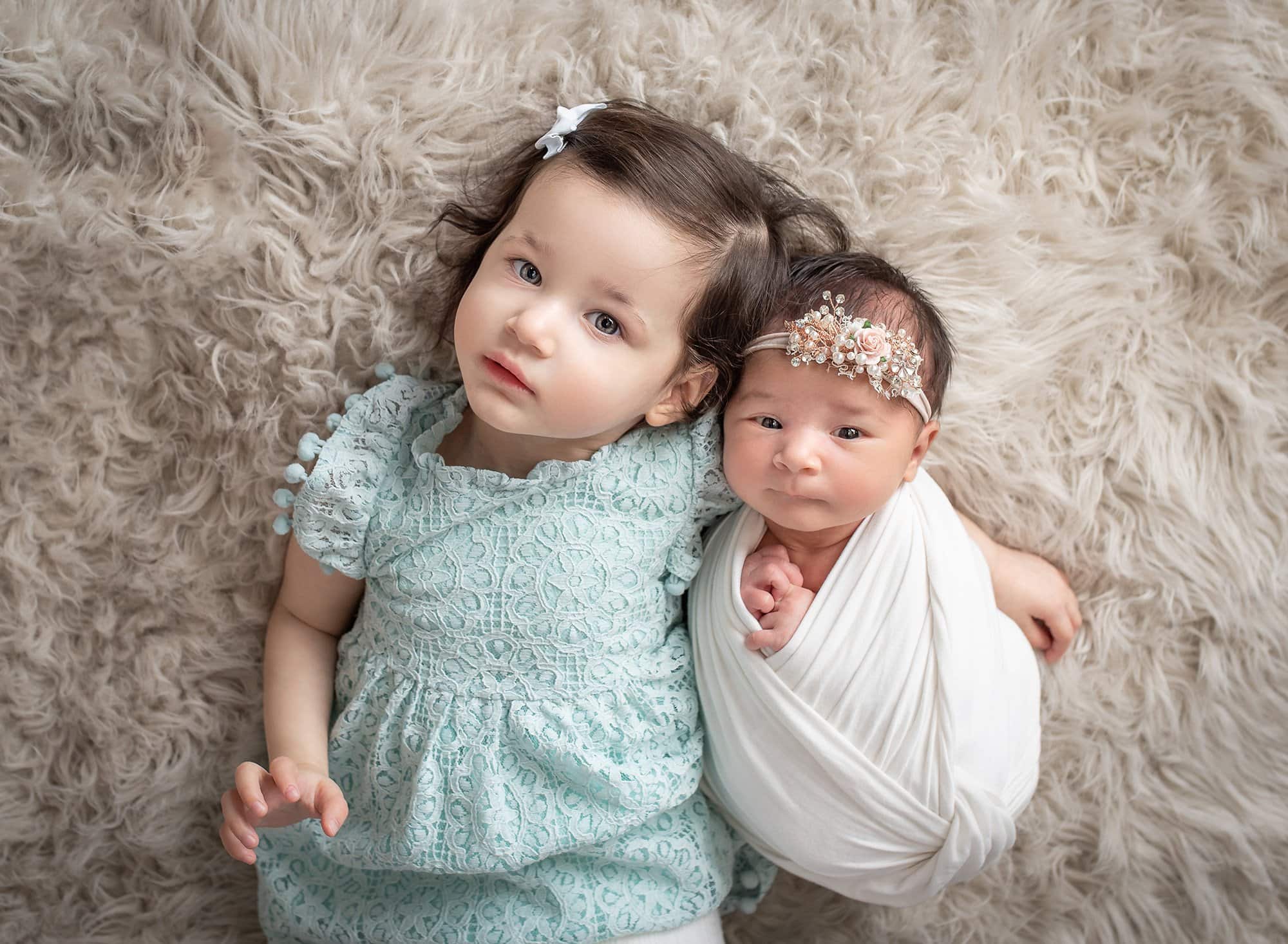 brunette young girl in a baby blue lace dress laying with newborn sister swaddled in white on fluffy cream backdrop