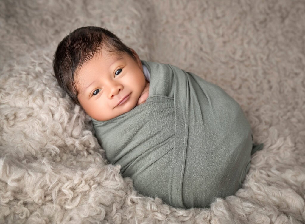 Awake newborn wrapped in sage on soft textured blanket, looking at camera