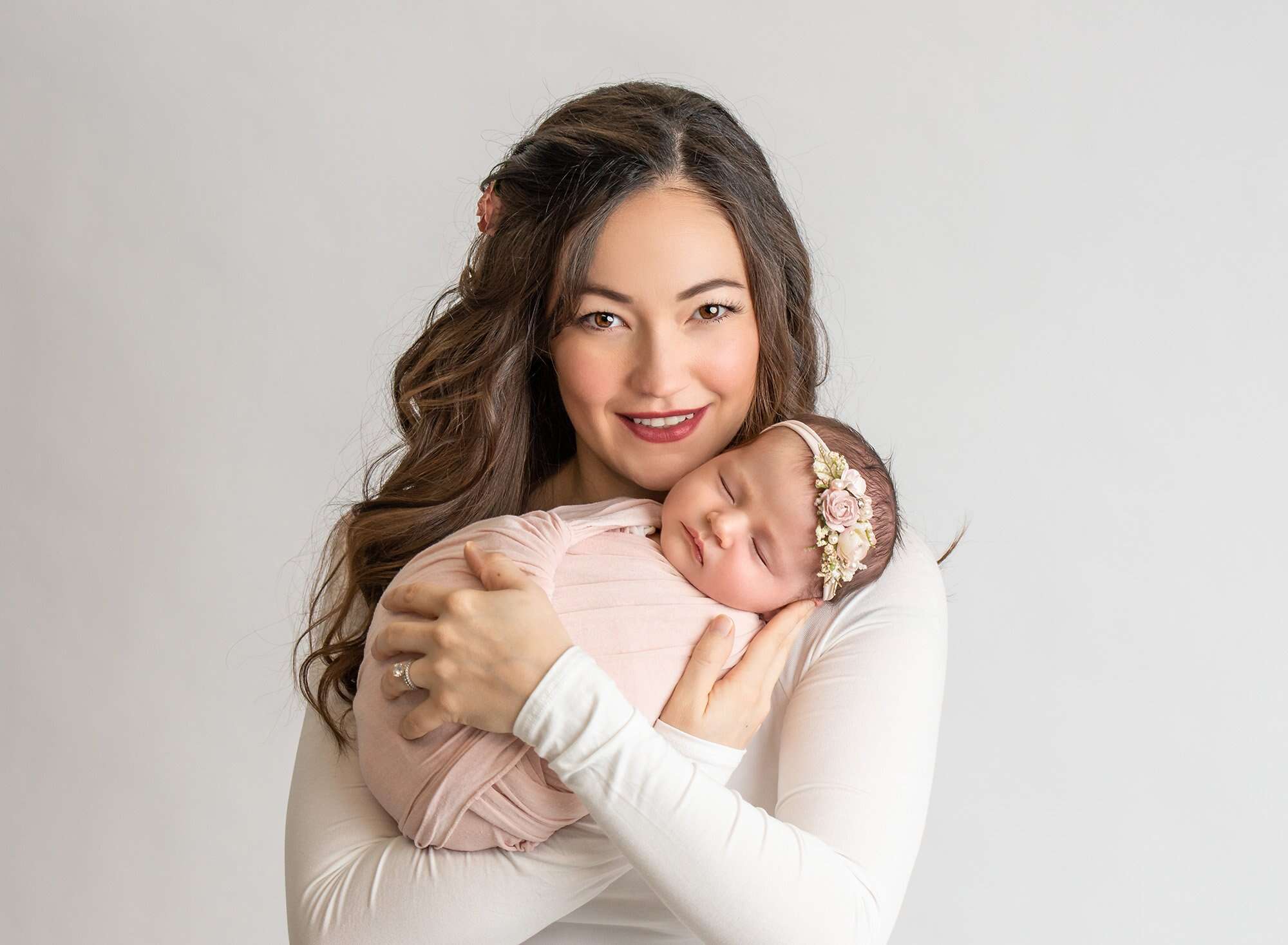 mother dressed in white on white backdrop cradling newborn baby girl swaddled in pink with floral headband