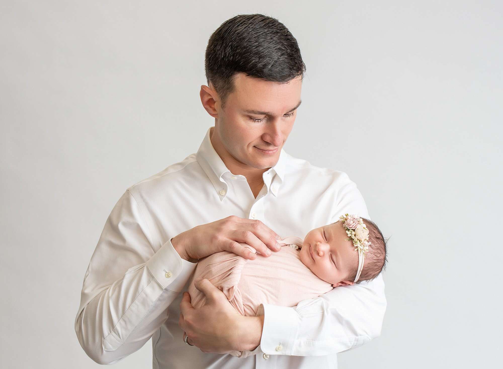 father in white looking down at his newborn baby girl swaddled in pink wearing floral headband