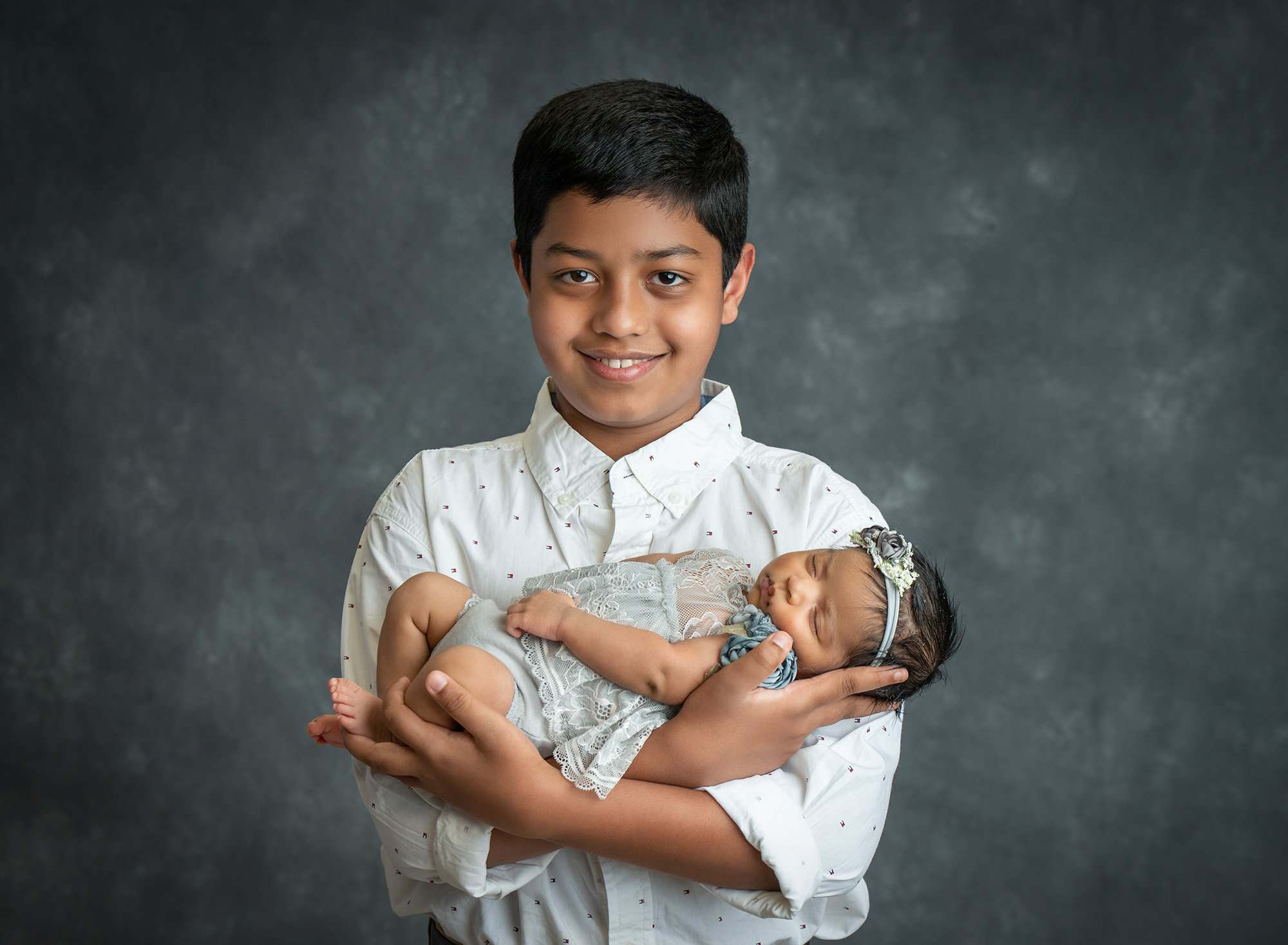 older brother proud to be holding asleep newborn baby sister dressed in a grey floral dress