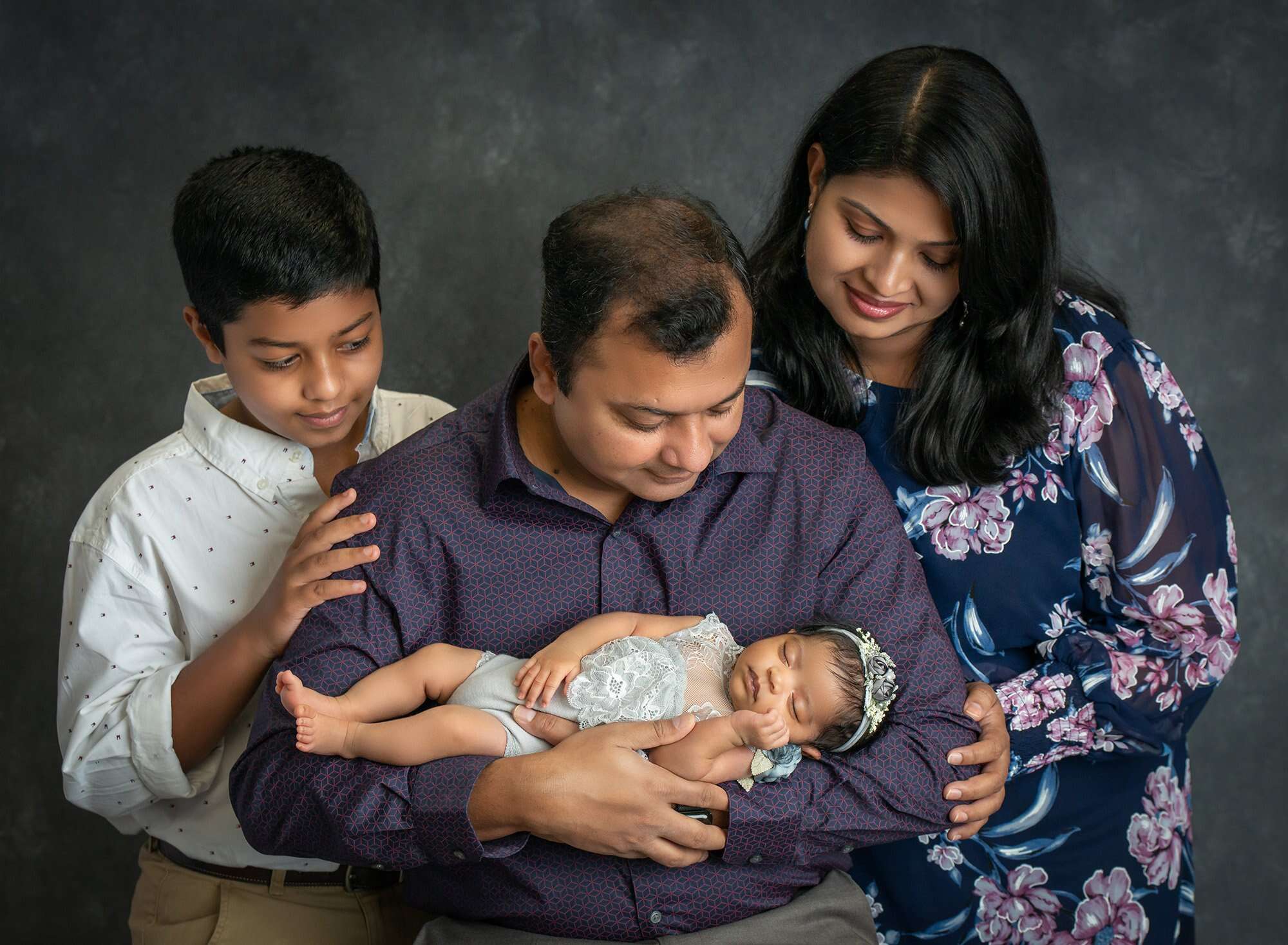 new parents and older son looking down at asleep newborn baby girl wearing floral grey dress