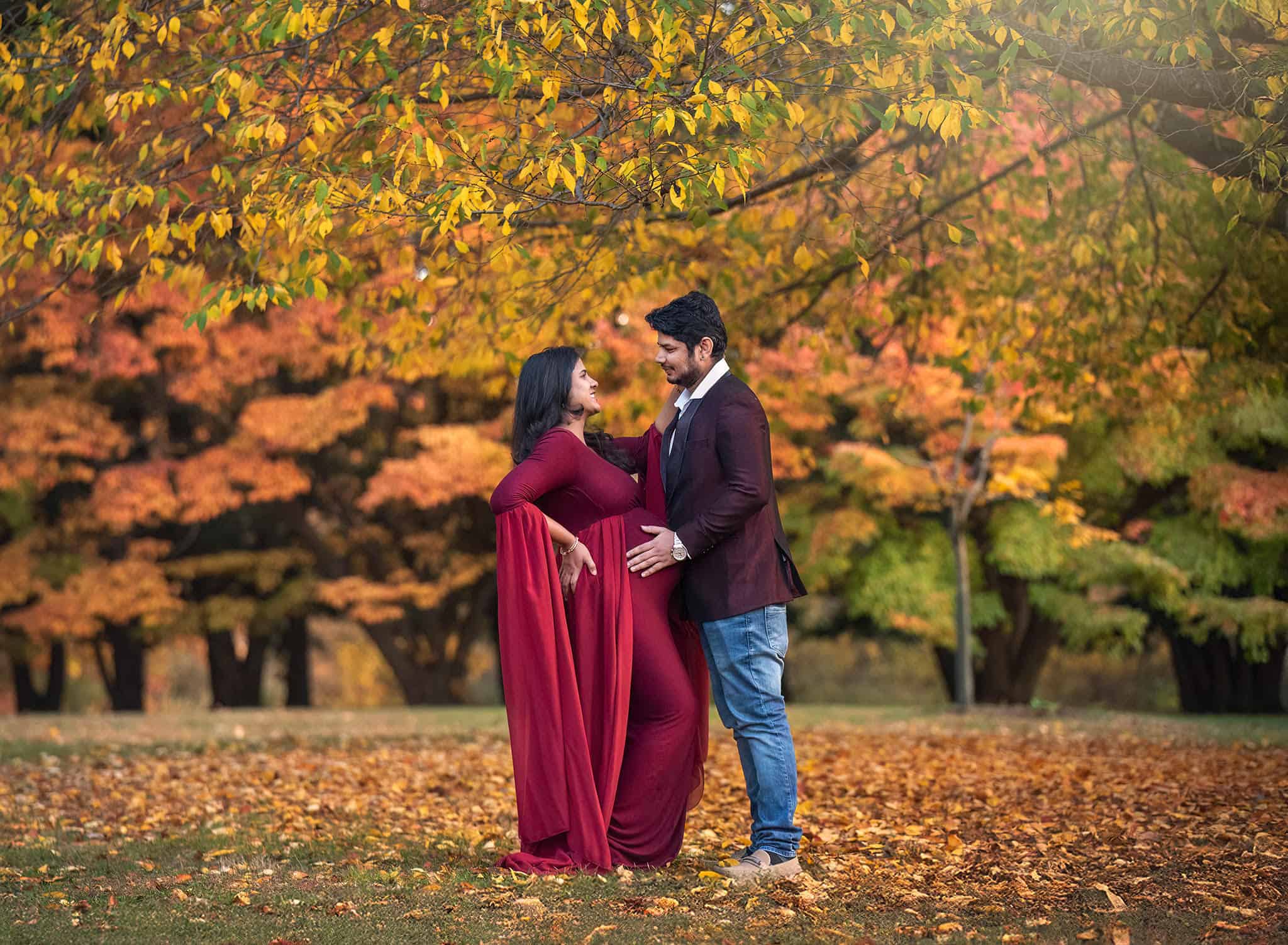 Pregnant couple in cranberry gown embracing baby belly in autumn landscape