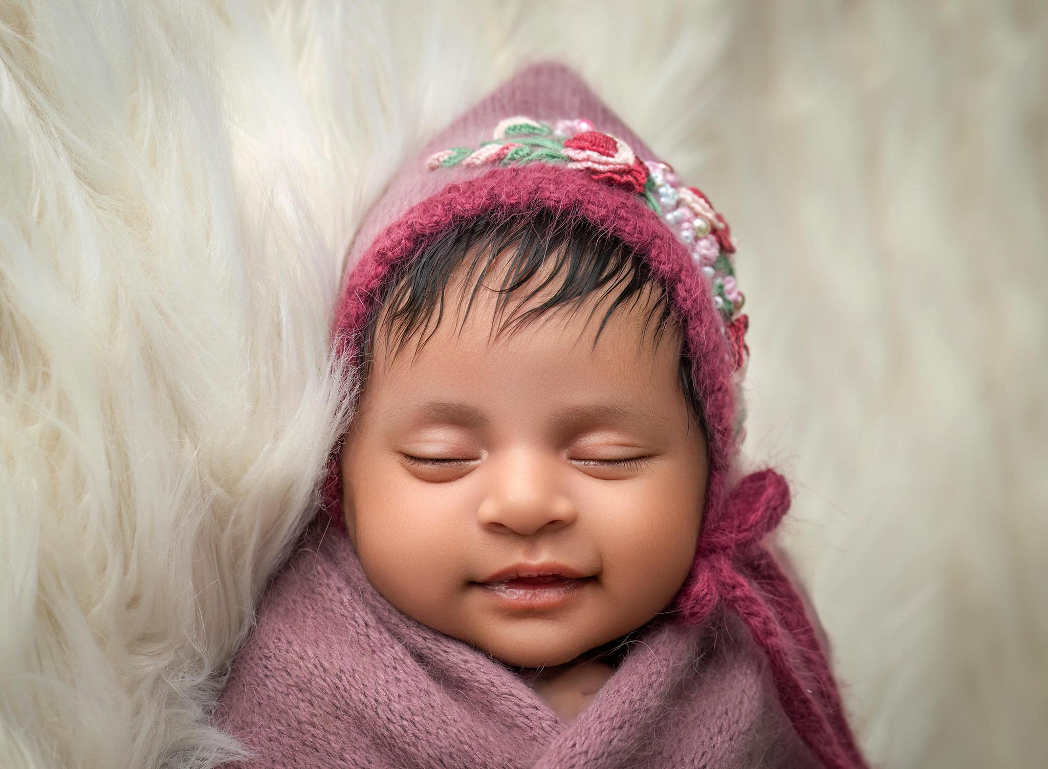newborn baby girl sleeping on cream furry blanket wearing a mauve wrap and bonnet