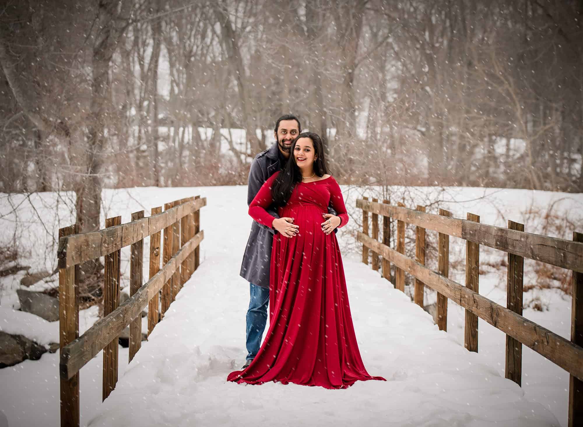 couples maternity photo with red maternity dress on a snowy bridge with falling snow