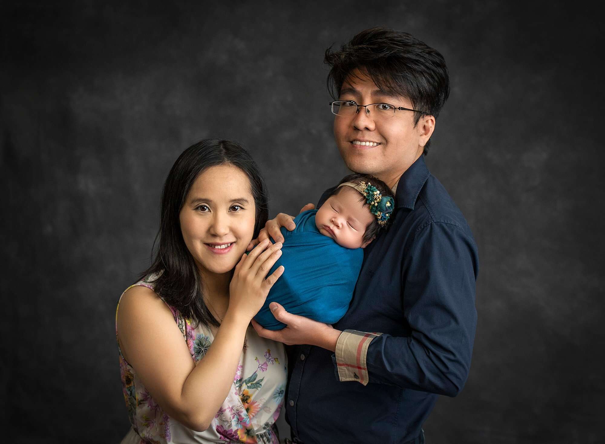 new parents holding sleeping newborn baby girl swaddled in teal wrap on dark grey background