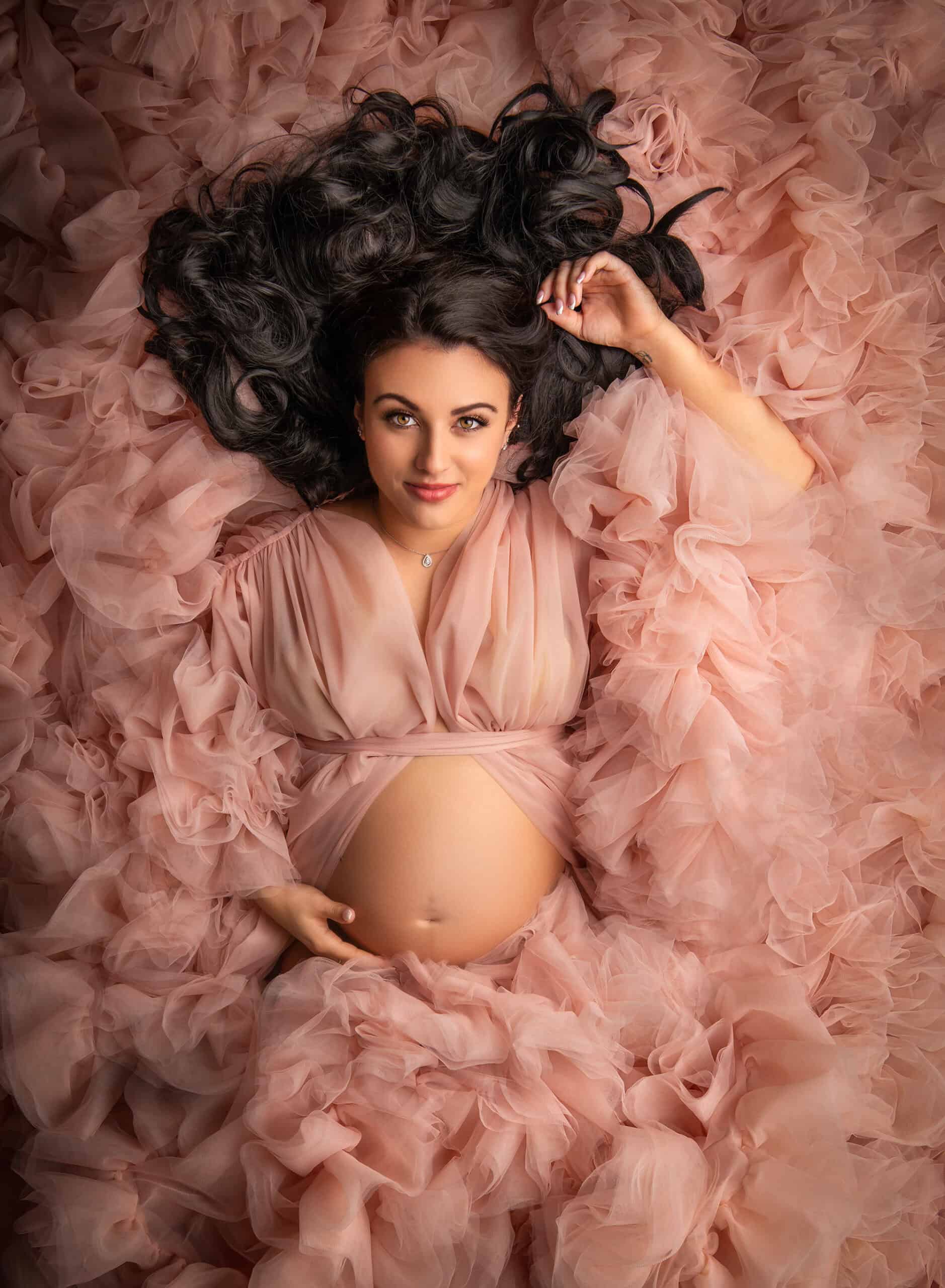 Maternity and Newborn Photography Mom in a pink fluffy gown, surrounded by ruffles, with her tummy showing.